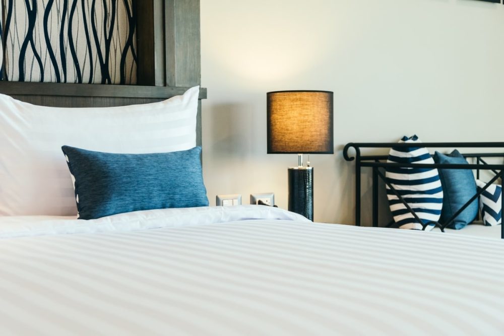 Close-up view of a bed in a staged home for sale, accessorised with carefully chosen cushions in shades of white and blue, adding a splash of colour and comfort. A striking piece of abstract artwork hangs above the bed, complementing the modern aesthetic. Warm lighting from a bedside lamp casts a cozy glow, illustrating the importance of accessories and artwork in staging a home for sale.