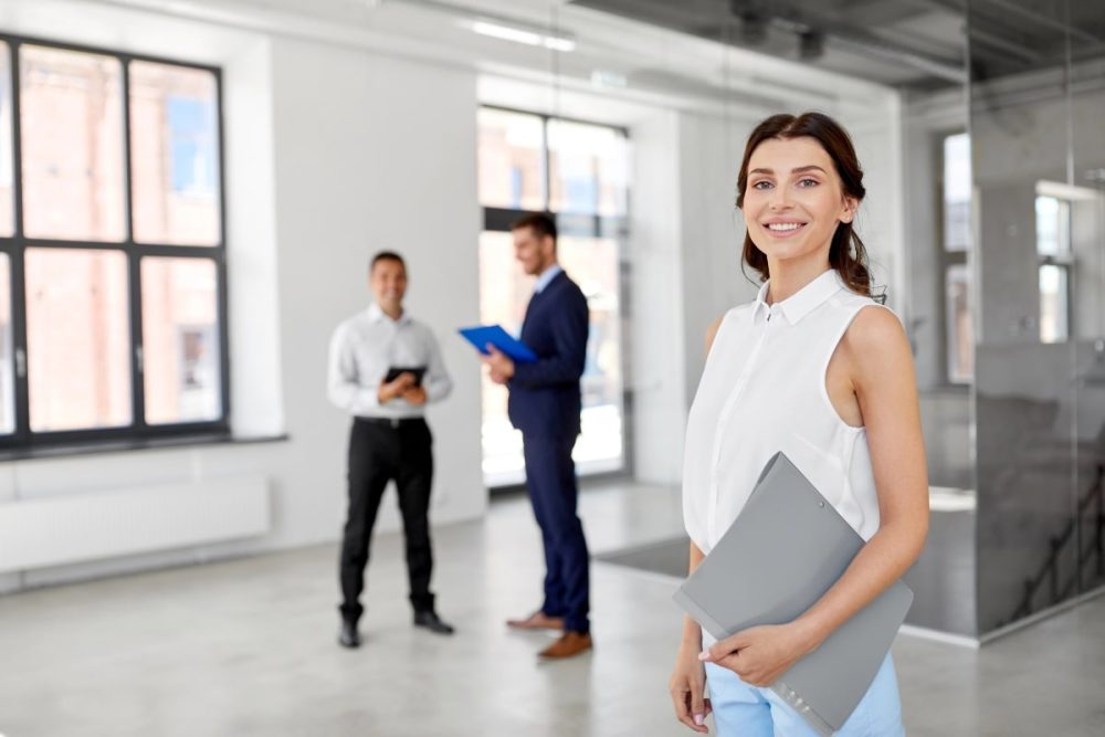 Smiling woman holding a folder in a modern office space with two colleagues in the background.