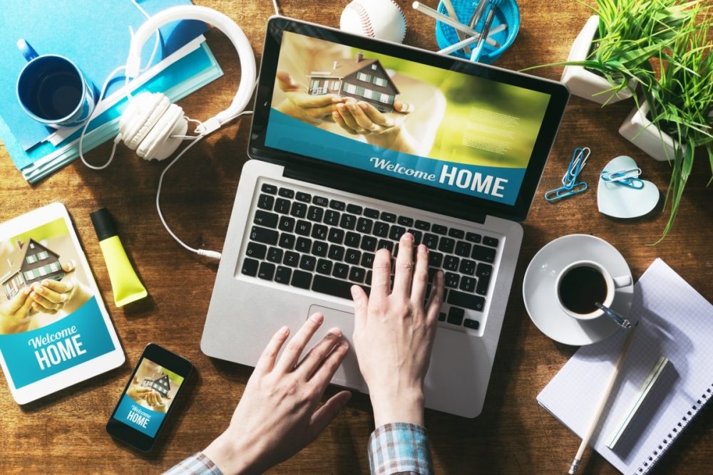 A person's hands typing on a laptop with the screen displaying a 'Welcome HOME' message, representing the research phase of buying a second house while renting out the first.