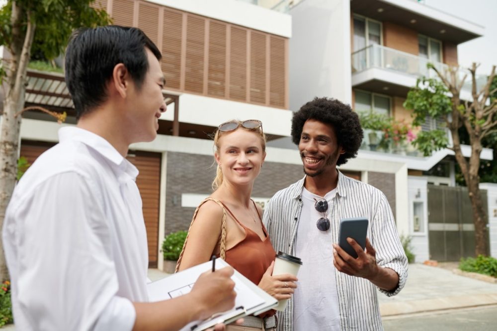 Prospective homebuyers talking to a real estate agent outdoors, showing enthusiasm while holding documents and a smartphone.