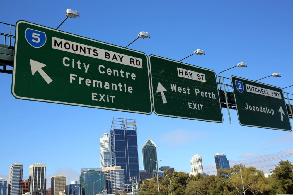 Freeway signs in Perth, Western Australia, showing exits for City Centre, West Perth, and Joondalup with the city skyline in the background.