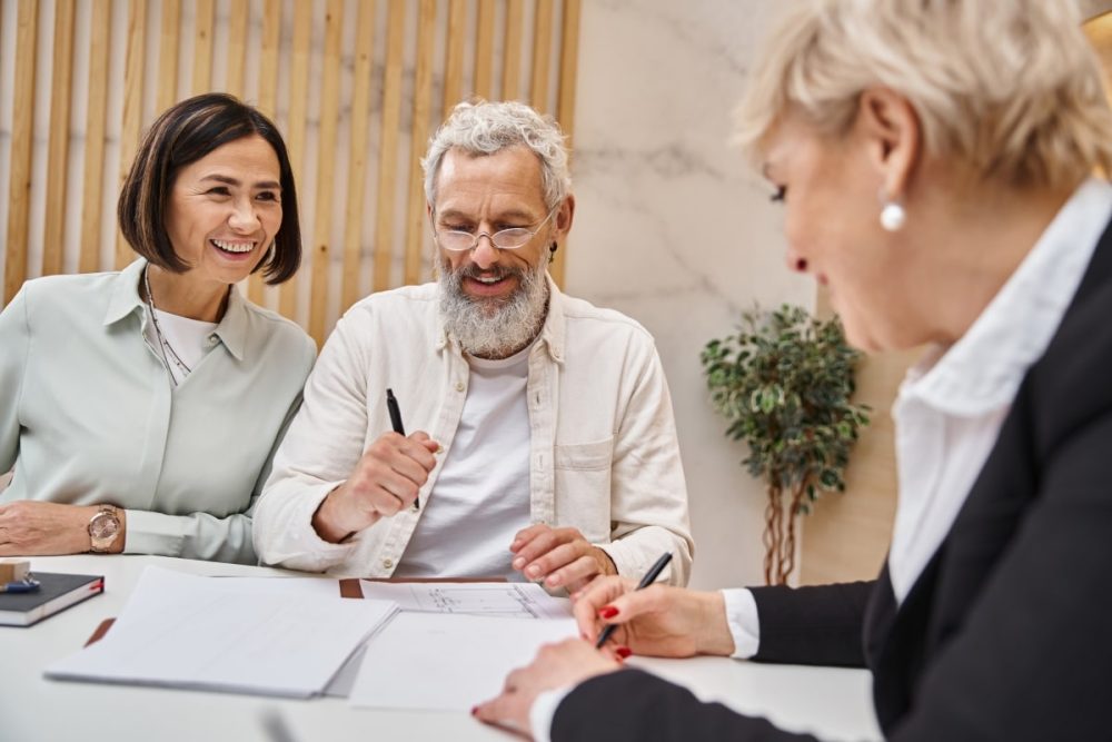 A mature couple smiling while signing documents, with a real estate agent, capturing a moment of closing and negotiations in the house selling journey.