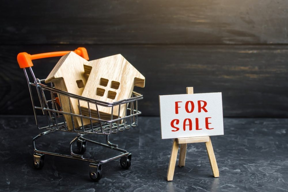 Shopping for a second house illustrated by wooden home miniatures in a shopping basket next to a 'For Sale' display, indicating the considerations in acquiring an additional home.